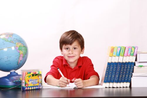 Ein lächelnder Junge im roten Shirt sitzt an einem Tisch und schreibt mit einem Stift in ein Heft, daneben stehen ein Globus und Bücher