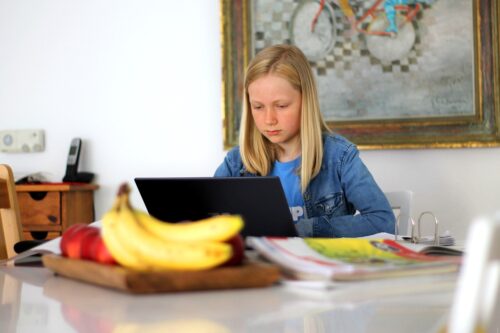 Ein Mädchen mit langen blonden Haaren sitzt zu Hause an einem Tisch und lernt konzentriert am Laptop